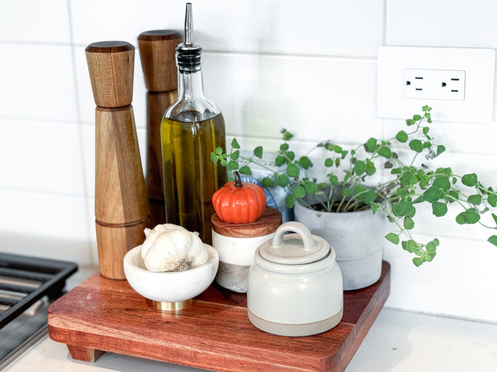 Salt and pepper grinders, olive oil cruet, salt cellar, fresh garlic, and fresh potted oregano sitting on a cutting board next to a stove