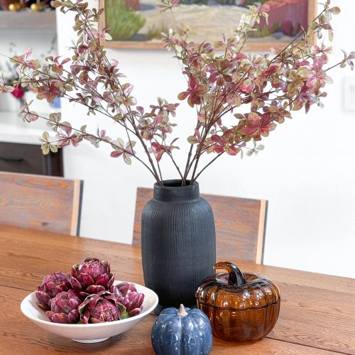 Fall stems in a black vase sitting on a dining table next to glass pumpkin decor