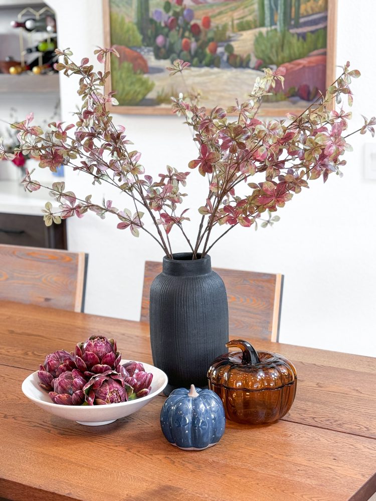 Fall stems in a black vase sitting on a dining table next to glass pumpkin decor