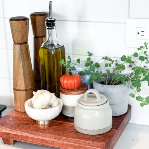 Salt and pepper grinders, olive oil cruet, salt cellar, fresh garlic, and fresh potted oregano sitting on a cutting board next to a stove