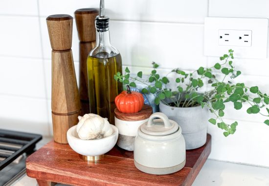 Salt and pepper grinders, olive oil cruet, salt cellar, fresh garlic, and fresh potted oregano sitting on a cutting board next to a stove